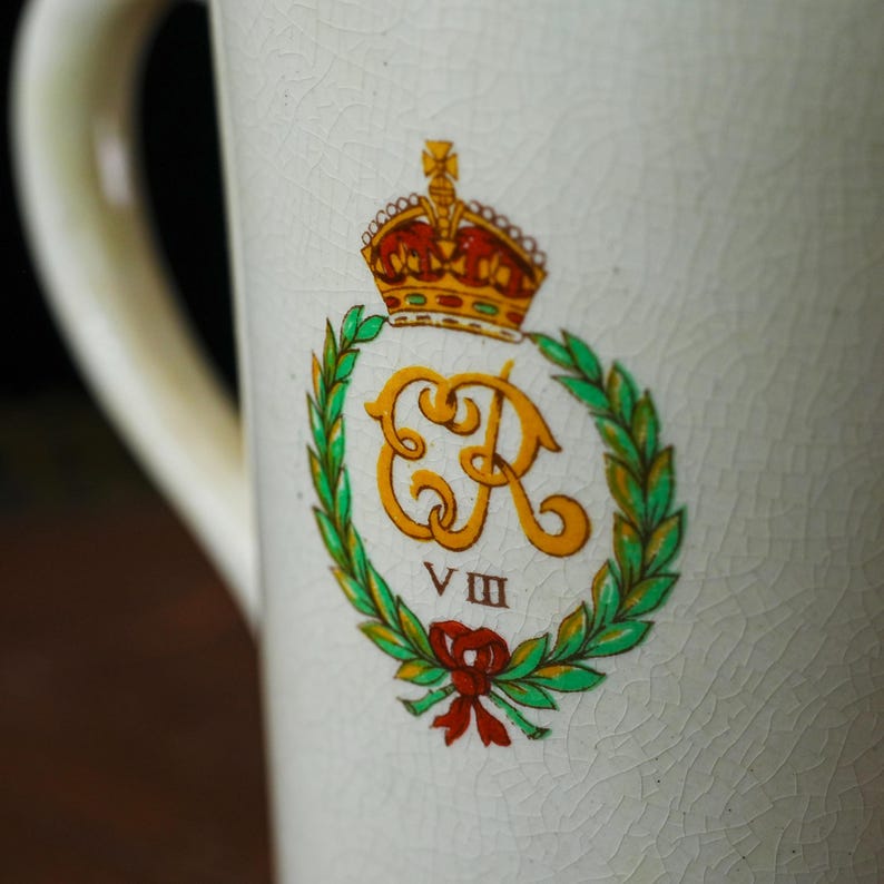 Puede incluir: Taza de cer&aacute;mica color crema con asa, con un emblema real. El emblema incluye una corona dorada, las letras "ER" en dorado, una corona de laurel verde y un lazo rojo. El n&uacute;mero romano "VIII" est&aacute; debajo.