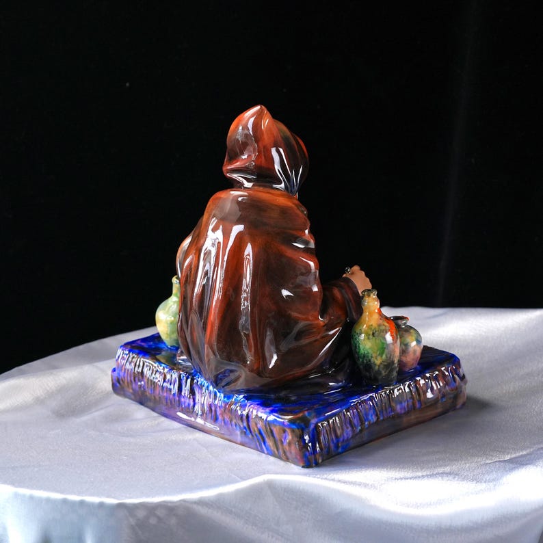 May include: A ceramic figurine depicting a robed figure, seen from behind, seated on a blue and brown base. The figure wears a brown hooded robe. Several small, colourful vases are arranged around the figure.
