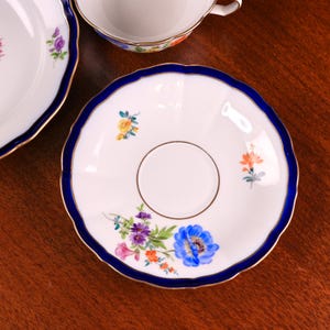 Puede incluir: Platillo de porcelana blanca con borde festoneado azul y ribete dorado. El platillo presenta un dise&ntilde;o floral con flores azules, moradas, naranjas y amarillas. En el fondo se ve una taza de t&eacute; a juego.