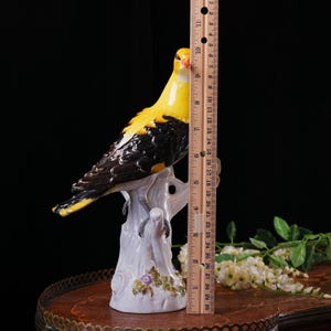 May include: A ceramic bird figurine, predominantly yellow and black, perched on a white tree branch. The bird's beak is orange, and the base features purple flowers. The figurine measures approximately 10 inches in height.