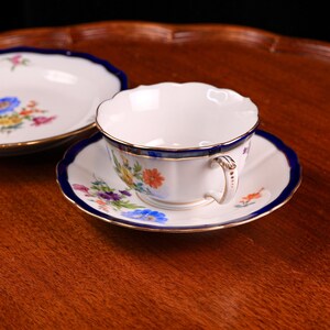 Puede incluir: Un juego de taza y platillo blancos con borde azul marino y ribete dorado. La taza y el platillo est&aacute;n decorados con coloridos estampados florales. El juego est&aacute; sobre una bandeja de madera.