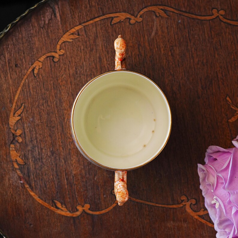 Puede incluir: Vista a&eacute;rea de una taza de t&eacute; vintage con interior crema y borde dorado. El asa y el exterior presentan un dise&ntilde;o moteado naranja y blanco. La taza se encuentra sobre una superficie de madera oscura con patrones incrustados, junto a una flor rosa.