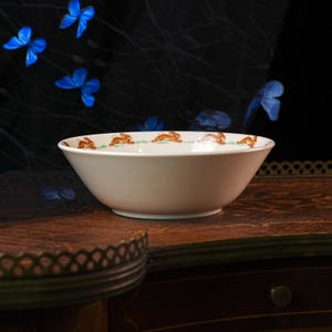 May include: A white ceramic bowl featuring a repeating pattern of brown rabbits running across a thin green grassy line along the interior rim. The bowl has a smooth, rounded shape with a slightly flared edge and a clean, minimalist white finish.