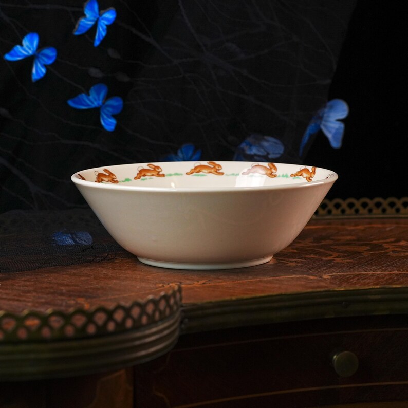 May include: A white ceramic bowl featuring a repeating pattern of brown rabbits running across a thin green grassy line along the interior rim. The bowl has a smooth, rounded shape with a slightly flared edge and a clean, minimalist white finish.