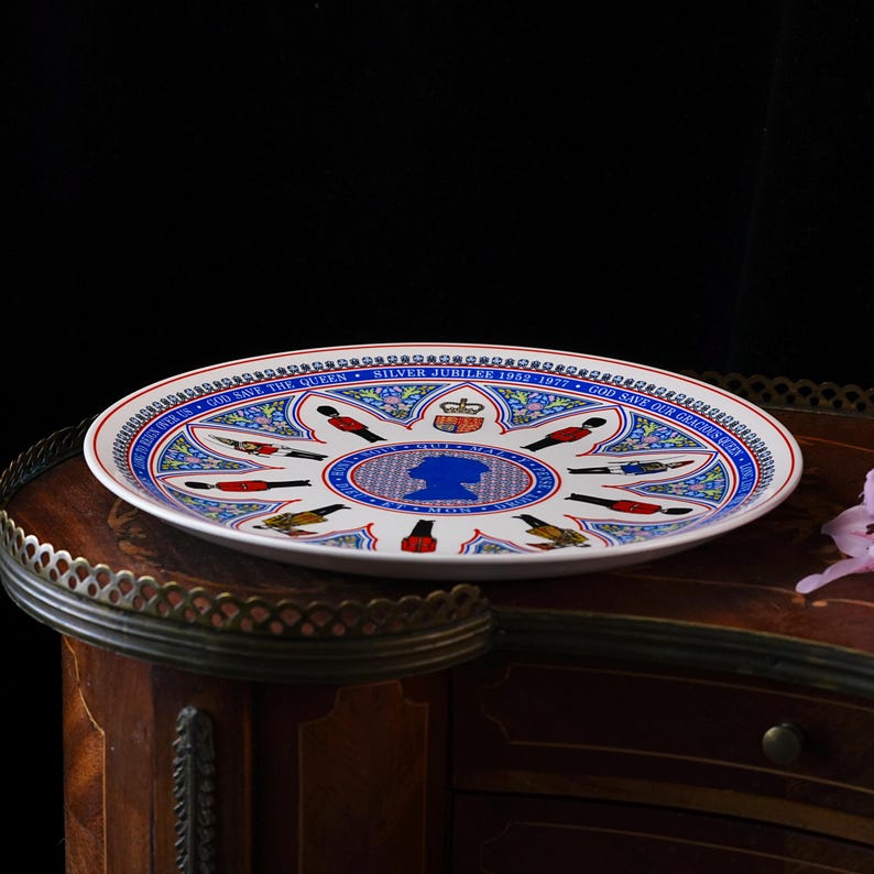 Puede incluir: Un plato conmemorativo de cer&aacute;mica blanca con una silueta azul de la Reina en el centro, rodeado de ilustraciones de guardias reales y motivos florales. El texto del borde dice Silver Jubilee 1952-1977 y God Save The Queen.