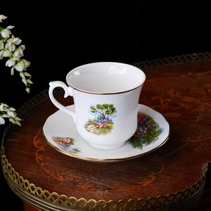 May include: A white teacup and saucer set with a gold rim. The cup and saucer feature a colorful landscape design with a tree, flowers, and a stone wall. The set is placed on a dark wooden surface.