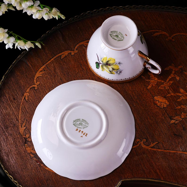 Peut inclure: Service &agrave; th&eacute; Royal Tuscan en porcelaine blanche avec un motif floral jaune et des bordures dor&eacute;es. La soucoupe porte l'inscription "Royal Tuscan" et le num&eacute;ro "D2962". L'ensemble est pr&eacute;sent&eacute; sur une table en bois.