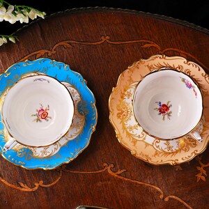 Op de afbeelding: Twee vintage kopjes en schotels. De ene set is lichtblauw met gouden randen, de andere is perzik met goud. Beide hebben bloemmotieven in het midden van de kop. De kopjes en schotels staan op een donker houten oppervlak.