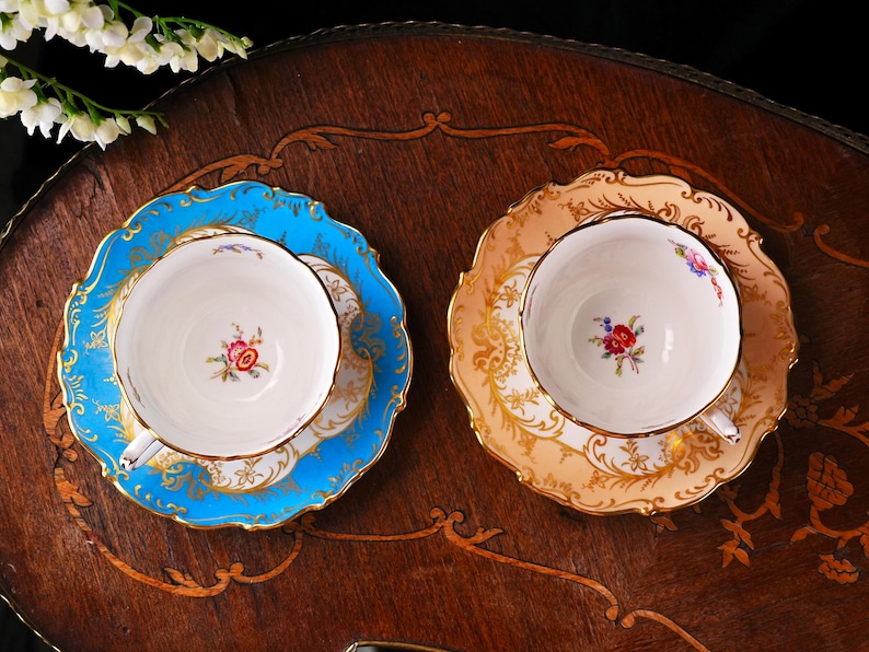 Op de afbeelding: Twee vintage kopjes en schotels. De ene set is lichtblauw met gouden randen, de andere is perzik met goud. Beide hebben bloemmotieven in het midden van de kop. De kopjes en schotels staan op een donker houten oppervlak.