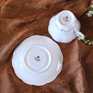 May include: A white teacup and saucer set with a scalloped edge. The teacup is upside down, revealing the Royal Albert stamp. The saucer is also white with a scalloped edge and the Royal Albert stamp. The set is on a brown patterned surface.
