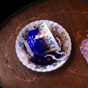 May include: A blue and gold teacup and saucer set. The teacup is a deep blue with gold floral detailing, and the saucer is white with gold accents. The set is placed on a wooden surface.