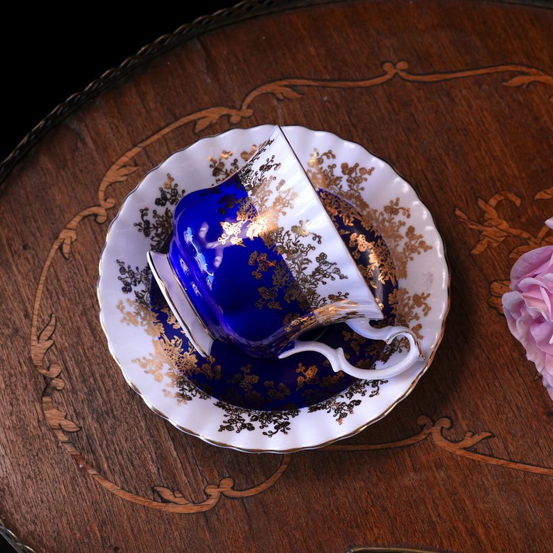 May include: A blue and gold teacup and saucer set. The teacup is a deep blue with gold floral detailing, and the saucer is white with gold accents. The set is placed on a wooden surface.