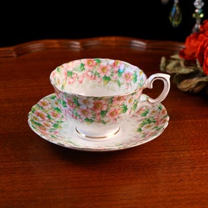 May include: A floral patterned teacup and saucer. The cup and saucer feature a design of pink and green flowers on a white base. The cup has a curved handle and a gold trim. The saucer has a scalloped edge, typical of vintage china.