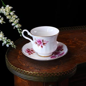 Op de afbeelding: Een witte kop en schotel set met een bloemen rozen design in roze en bruin. De kop en schotel hebben een gouden rand. De set staat op een donkere houten tafel met een decoratieve rand. Een takje witte bloemen staat op de achtergrond.