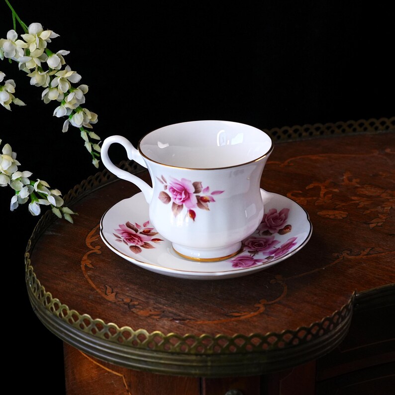 Op de afbeelding: Een witte kop en schotel set met een bloemen rozen design in roze en bruin. De kop en schotel hebben een gouden rand. De set staat op een donkere houten tafel met een decoratieve rand. Een takje witte bloemen staat op de achtergrond.