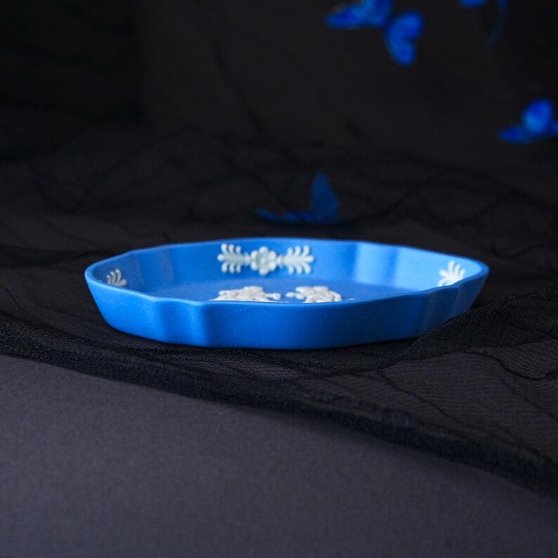 May include: A bright blue shallow dish with a scalloped edge and white relief detailing. The interior features white embossed floral and foliate motifs along the rim and a central white relief design of two figures in a classic style.