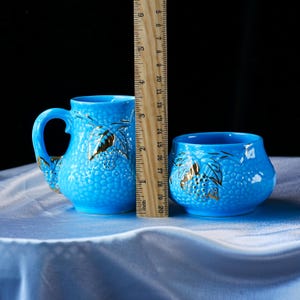 Op de afbeelding: Een felblauwe keramische mok en een bijpassende kom, beide met een getextureerd oppervlak en goudkleurige druivenbladaccenten. Een houten liniaal staat ertussen en toont dat de mok ongeveer 11,5 cm hoog is.