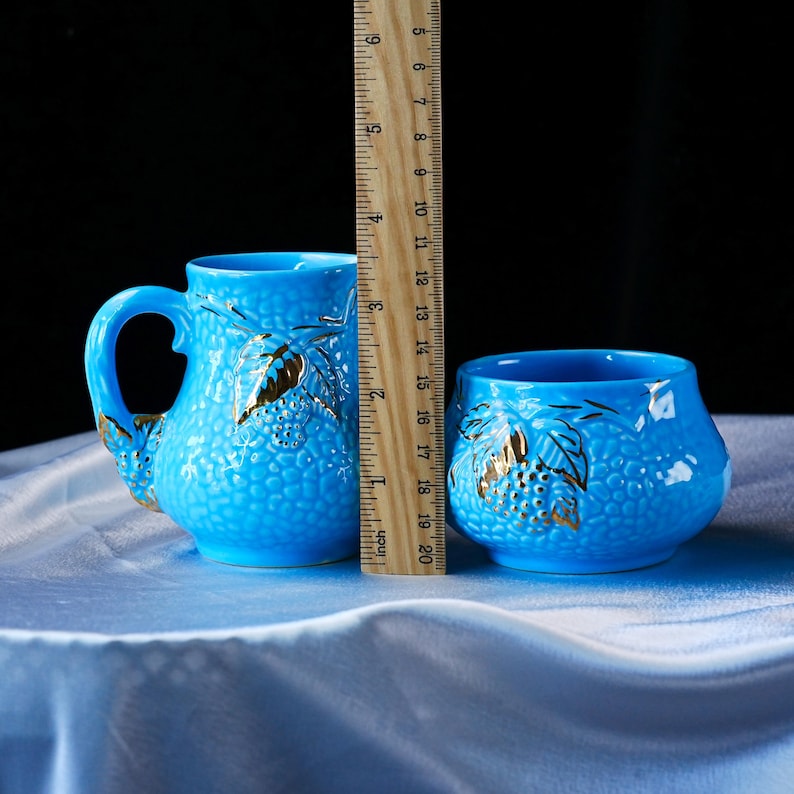 Op de afbeelding: Een felblauwe keramische mok en een bijpassende kom, beide met een getextureerd oppervlak en goudkleurige druivenbladaccenten. Een houten liniaal staat ertussen en toont dat de mok ongeveer 11,5 cm hoog is.