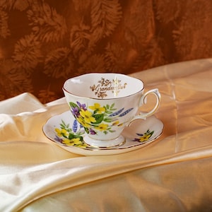 K&ouml;nnte beinhalten: Ein wei&szlig;es Tassen- und Untertassenset mit einem Blumendesign mit gelben und lila Blumen. Die Tasse tr&auml;gt den Schriftzug "Grandmother" in goldener Schrift. Das Set hat goldene Verzierungen und liegt auf einer Satin-Oberfl&auml;che.