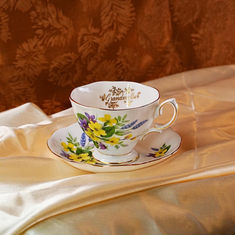 K&ouml;nnte beinhalten: Ein wei&szlig;es Tassen- und Untertassenset mit einem Blumendesign mit gelben und lila Blumen. Die Tasse tr&auml;gt den Schriftzug "Grandmother" in goldener Schrift. Das Set hat goldene Verzierungen und liegt auf einer Satin-Oberfl&auml;che.