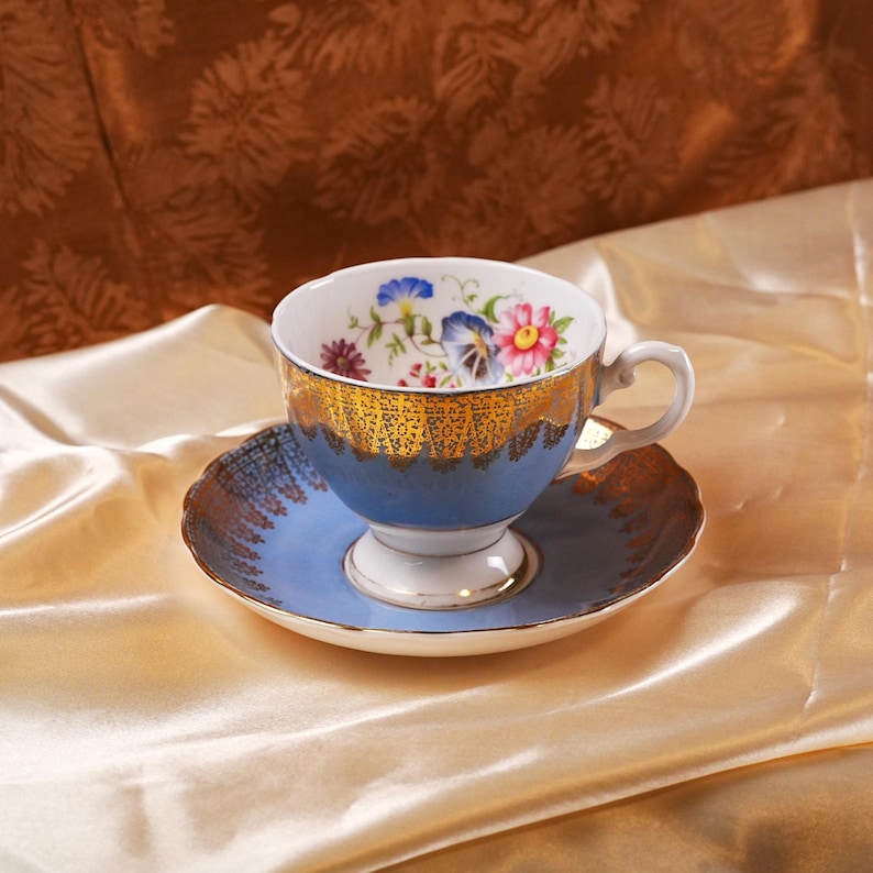 Op de afbeelding: Een blauwe theekop en schotel set met gouden details en bloemmotieven. De theekop heeft een witte binnenkant met kleurrijke bloemen en een blauwe buitenkant met gouden accenten. De schotel is blauw met goud, wat een elegante uitstraling cre&euml;ert.