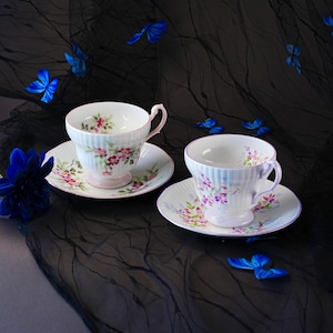 May include: Two white porcelain teacups and matching saucers featuring delicate pink and purple floral patterns with ribbed textures. The cups have curved handles and pedestal bases, decorated with small painted blossoms and green leaves.
