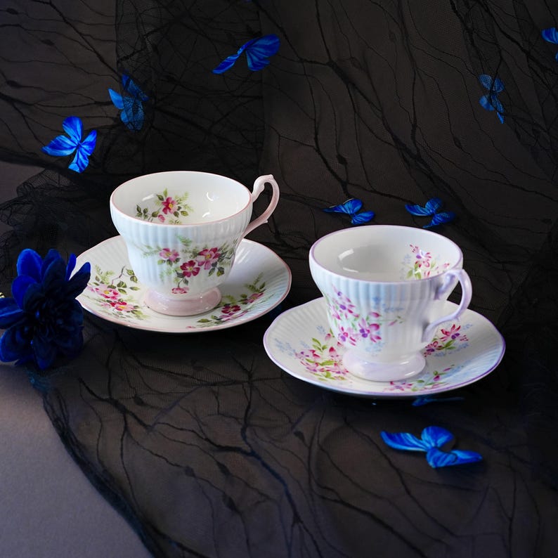 May include: Two white porcelain teacups and matching saucers featuring delicate pink and purple floral patterns with ribbed textures. The cups have curved handles and pedestal bases, decorated with small painted blossoms and green leaves.