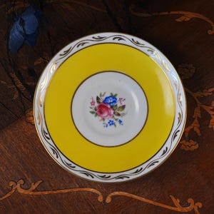 Op de afbeelding: Een rond porseleinen schoteltje met een felgele rand, een wit midden en een centraal bloemmotief met roze, blauwe en groene details. De rand is versierd met een goudkleurig filigraanpatroon en een dunne goudkleurige rand.