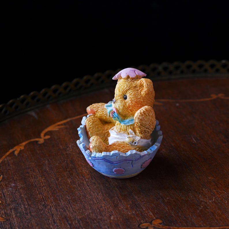 May include: A small, detailed figurine of a teddy bear seated in a blue and white decorative bowl. The bear is light brown with a pink hat and a blue scarf. The bowl has a floral design.
