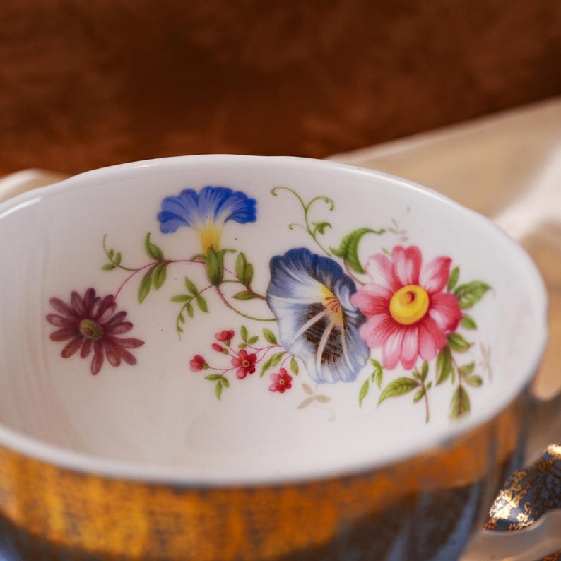 Op de afbeelding: Een witte keramische kom met een bloemenontwerp met blauwe, roze en paarse bloemen. De kom heeft een gouden rand en een blauw-gouden basis. De bloemen zijn gedetailleerd met groene bladeren en stelen.