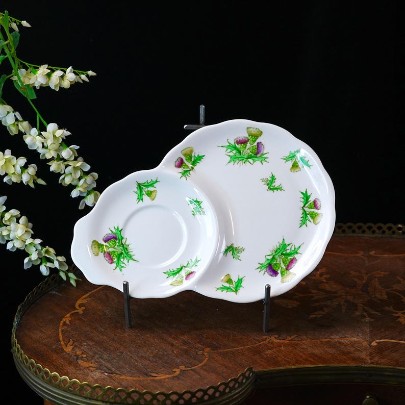 Puede incluir: Juego de plato y platillo de cer&aacute;mica blanca con dise&ntilde;o de cardo. El plato tiene una forma &uacute;nica con borde festoneado, y el platillo es redondo. El dise&ntilde;o de cardo presenta detalles en morado, verde y amarillo.
