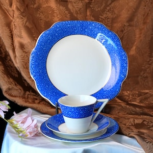 May include: A vintage blue and white china tea set. The set includes a large plate, a teacup, a saucer, and a smaller plate. The blue detailing has a speckled pattern. The teacup and saucer are white with a blue band. The large plate has a scalloped edge.