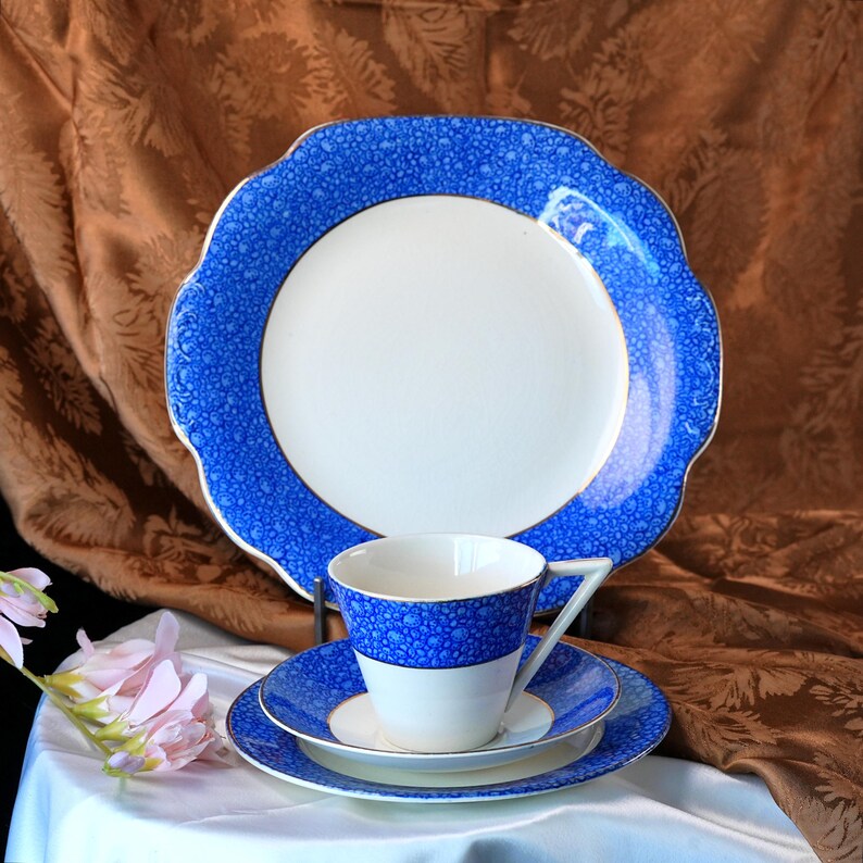May include: A vintage blue and white china tea set. The set includes a large plate, a teacup, a saucer, and a smaller plate. The blue detailing has a speckled pattern. The teacup and saucer are white with a blue band. The large plate has a scalloped edge.