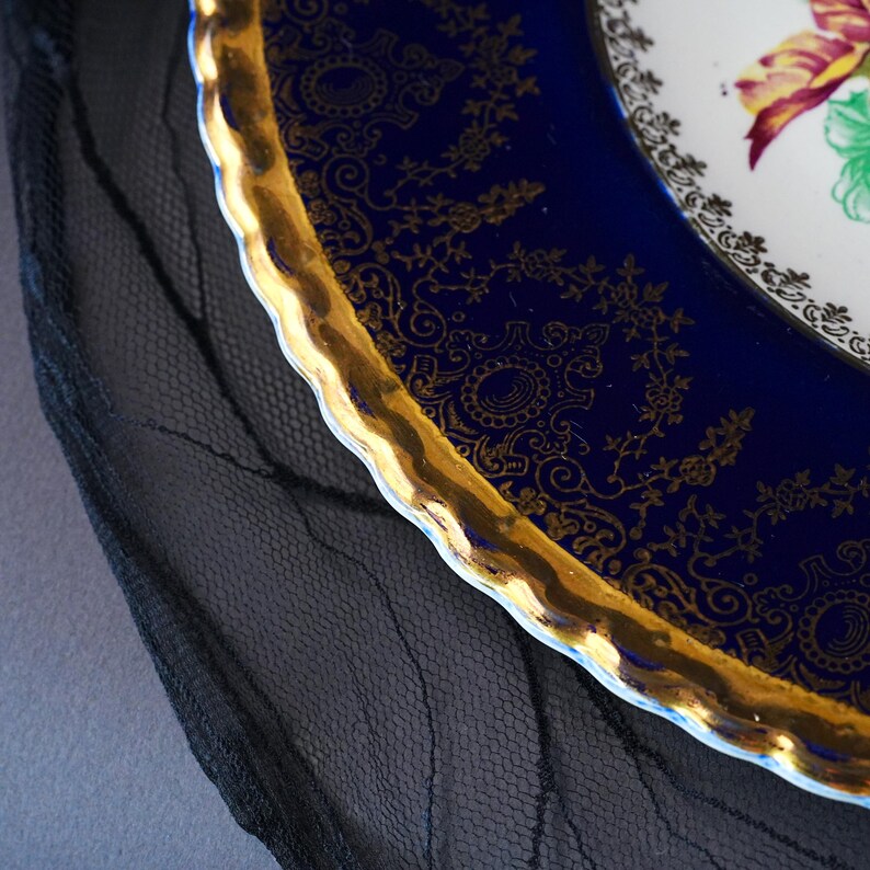 May include: A decorative plate featuring a deep cobalt blue border with intricate gold filigree patterns and a scalloped, gilded edge. The center of the plate displays a white surface with a partial floral design in shades of pink and green.