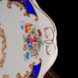 Puede incluir: Un plato de porcelana blanca con borde festoneado y ribete dorado. La superficie est&aacute; decorada con un ramo de flores en tonos rosa, azul, naranja y verde, enmarcado por paneles azul oscuro y complejos patrones de volutas doradas.