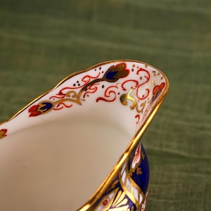 May include: Close-up of a white porcelain creamer with intricate hand-painted floral designs in red, blue, and gold. The rim is trimmed with gold, and the base is a deep blue. The creamer has a classic, elegant shape.
