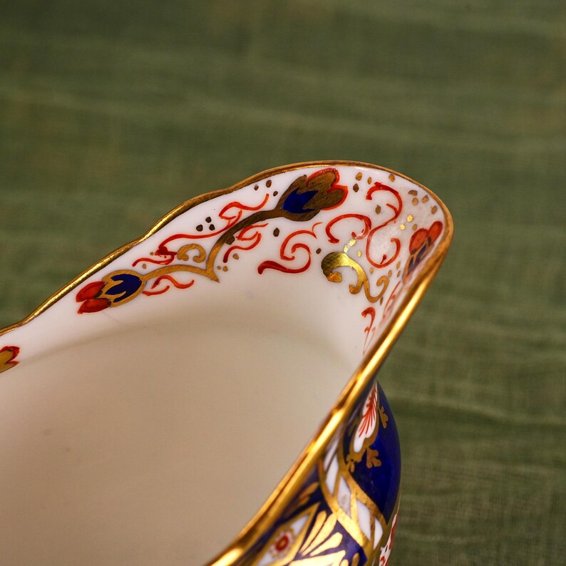 May include: Close-up of a white porcelain creamer with intricate hand-painted floral designs in red, blue, and gold. The rim is trimmed with gold, and the base is a deep blue. The creamer has a classic, elegant shape.