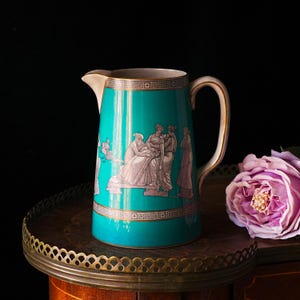 Puede incluir: Una jarra de cerámica turquesa con una escena clásica con figuras vestidas con telas drapeadas. La jarra tiene un borde y un asa dorados, y está colocada sobre una bandeja decorativa con una rosa rosa en el fondo.
