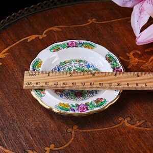 Puede incluir: Un peque&ntilde;o plato conmemorativo de cer&aacute;mica blanca con borde festoneado dorado, que presenta un borde floral colorido y el texto Coronation 2nd June 1953 Queen Elizabeth II. Una regla de madera muestra un di&aacute;metro de aproximadamente 11,5 cm.