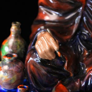 May include: A ceramic sculpture featuring a hand holding a paintbrush, with a red and blue glaze. Several small, colourful ceramic bottles are in the background. The artwork has a glossy finish against a black backdrop.