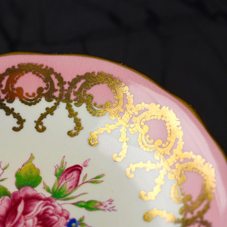 Puede incluir: Un platillo de porcelana con un borde rosa suave y filigrana dorada intrincada. El centro del platillo muestra un arreglo floral detallado con rosas de color rosa vibrante, hojas verdes y peque&ntilde;as flores azules sobre un fondo blanco.