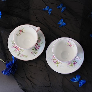 May include: Two white porcelain teacups and matching saucers featuring delicate floral patterns in pink, purple, and green. The saucers have a ribbed texture and thin pink rims. The set is arranged on a black fabric surface with blue butterfly accents.