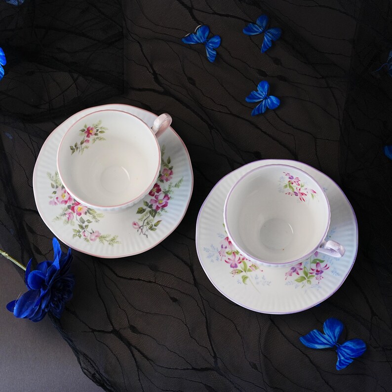 May include: Two white porcelain teacups and matching saucers featuring delicate floral patterns in pink, purple, and green. The saucers have a ribbed texture and thin pink rims. The set is arranged on a black fabric surface with blue butterfly accents.