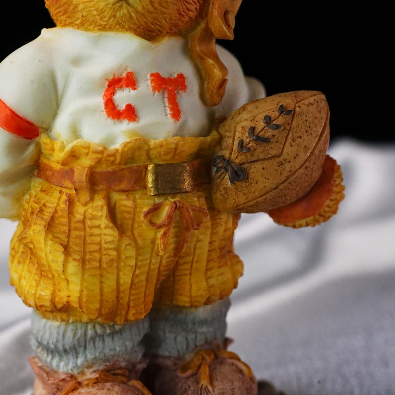 Op de afbeelding: Een beeldje van een beer met een wit shirt met oranje letters "CT" en een gele korte broek. De beer houdt een bruine voetbal vast met decoratieve bladeren. Het beeldje heeft een bruine riem en oranje accenten.