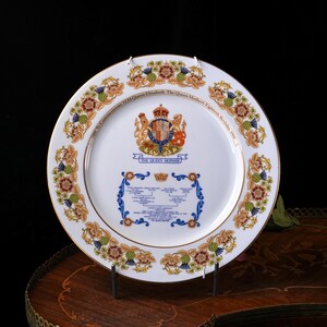 Pode incluir: Um prato comemorativo de porcelana branca com borda dourada, um bras&atilde;o real central e o texto To Commemorate HM Queen Elizabeth The Queen Mother's Eightieth Birthday 1980. A borda exibe motivos florais, incluindo flores de cardo.