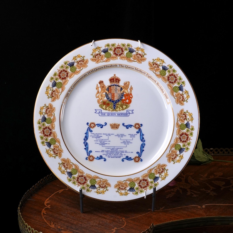 Pode incluir: Um prato comemorativo de porcelana branca com borda dourada, um bras&atilde;o real central e o texto To Commemorate HM Queen Elizabeth The Queen Mother's Eightieth Birthday 1980. A borda exibe motivos florais, incluindo flores de cardo.