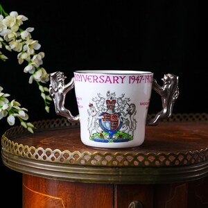 May include: A white ceramic mug with silver lion-shaped handles. The mug features a royal crest and the text "Anniversary 1947-1972" in purple. The mug is displayed on a wooden surface with a decorative brass edge.