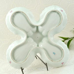 May include: A pale blue, four-sectioned serving dish with a distinctive clover shape. The dish features a scalloped edge and is supported by a metal stand. Small pink floral accents are visible on the rim. The dish is made of porcelain.