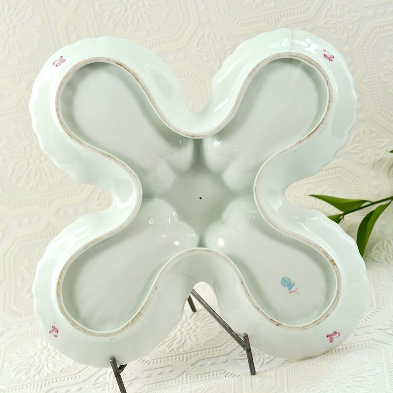 May include: A pale blue, four-sectioned serving dish with a distinctive clover shape. The dish features a scalloped edge and is supported by a metal stand. Small pink floral accents are visible on the rim. The dish is made of porcelain.