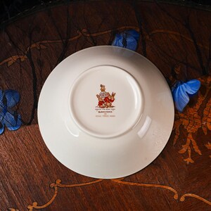May include: A white ceramic saucer featuring a central stamp with a crown, a group of rabbits, and the text English Fine Bone China Bunnykins Regd. Trade Mark. The underside of the saucer is smooth and circular with a raised foot rim.